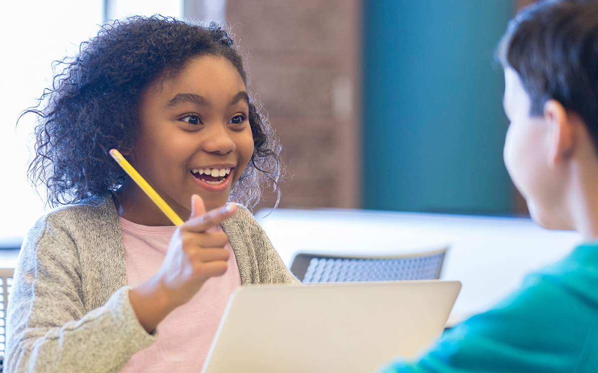 Photo of a young student sitting in front of a laptop and engaging with another student, who is seen from the back.