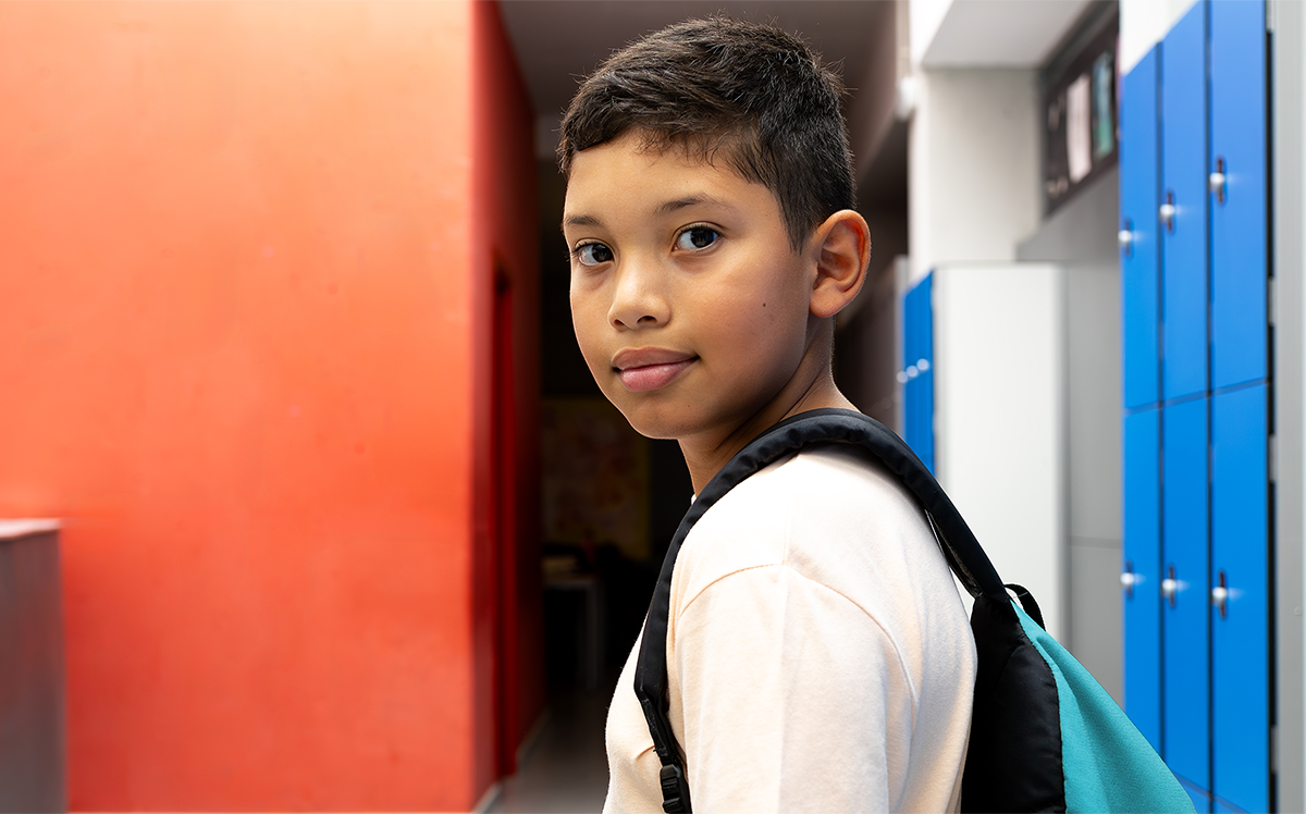 Photo of a young student standing in a colourful classroom and wearing a backpack on one shoulder, turning to face us.