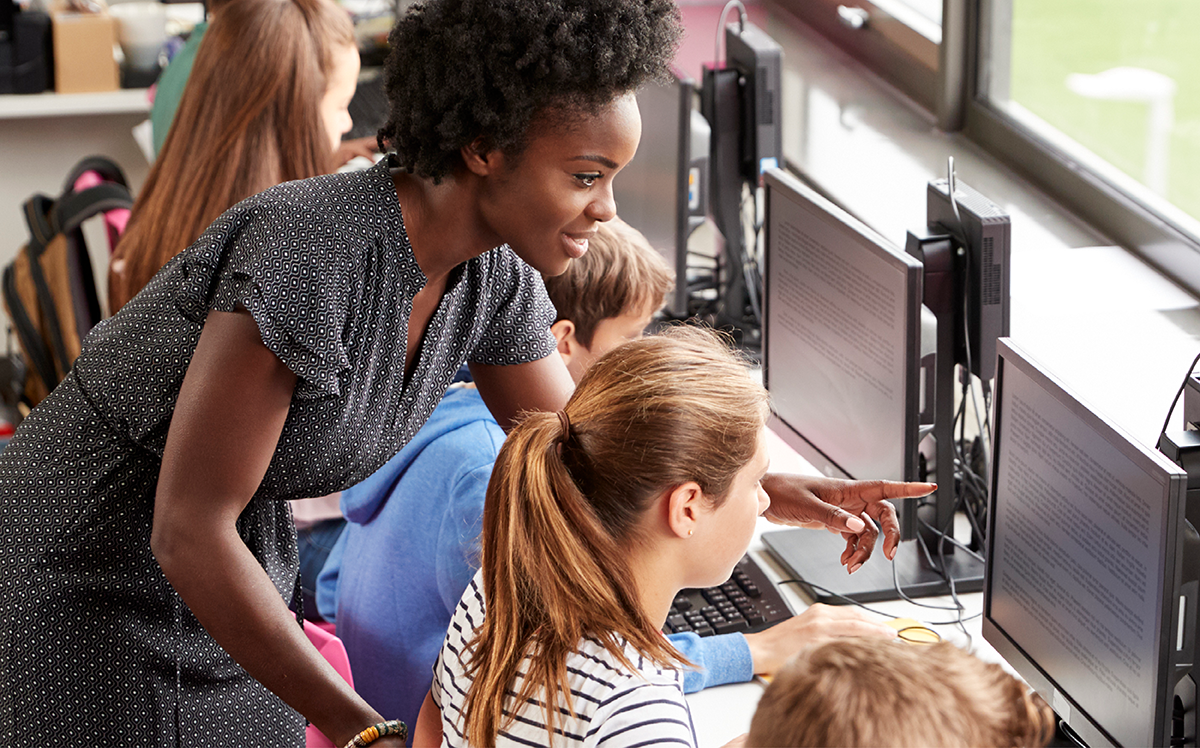 Photo of a teacher in a classroom where four students are working on computers; the teacher is helping one of the students.