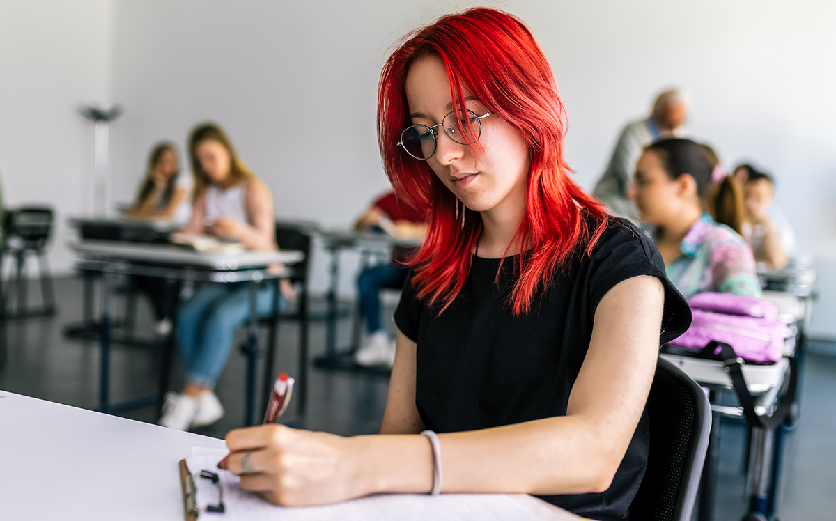 Photo of adolescents sitting in a classroom at individual desks, with a student in the foreground writing with a pen.