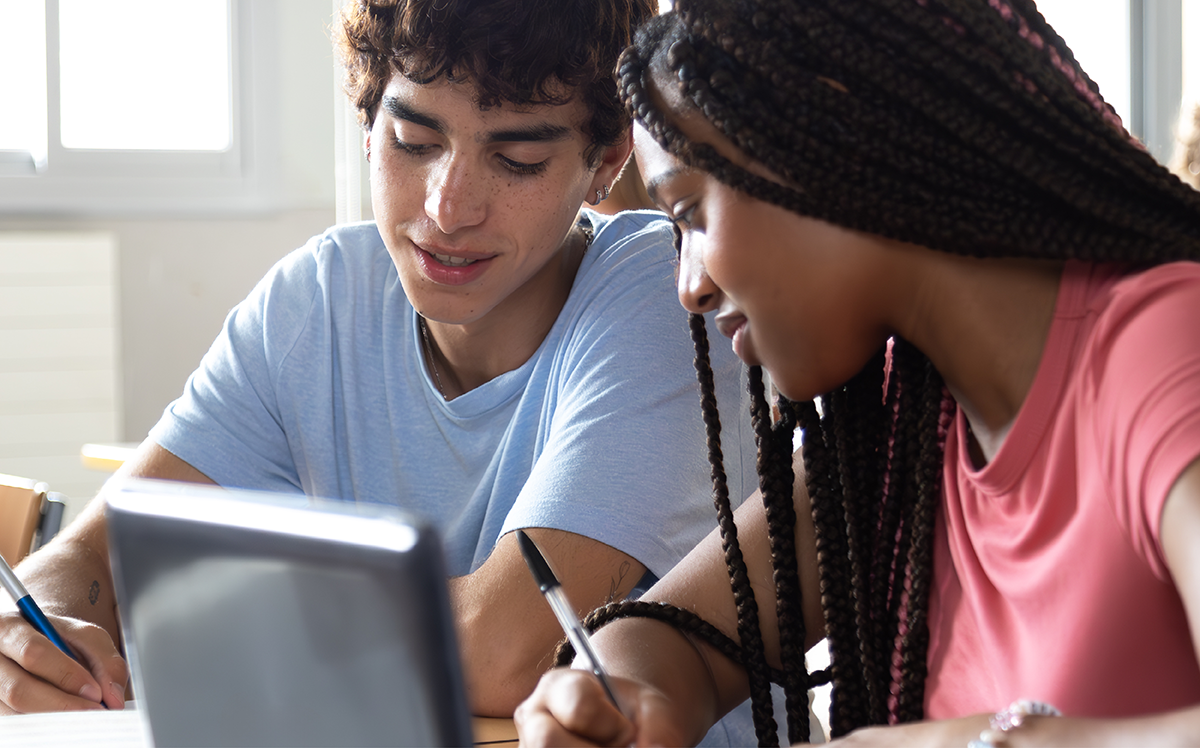 Photo of two adolescent students holding pens and working together, with a tablet on a stand in the foreground.